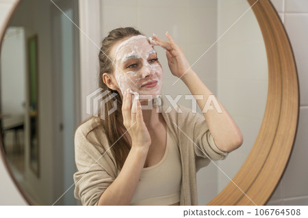 A young woman using an emollient facial moisturizer for facial skin care. Cosmetic procedures. A woman puts makeup on her face while relaxing in the bathroom 106764508
