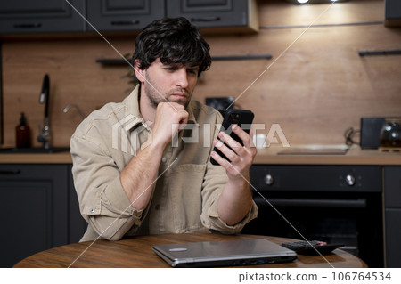 A young businessman sends messages on a smartphone while sitting at a table in the kitchen A young businessman sends messages on a smartphone while sitting at a table in the kitchen 106764534