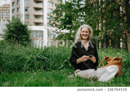 Portrait of beautiful woman with gray hair sitting on the grass in city park, using her smartphone. Portrait of beautiful woman with gray hair sitting on the grass in city park, using her smartphone. 106764538