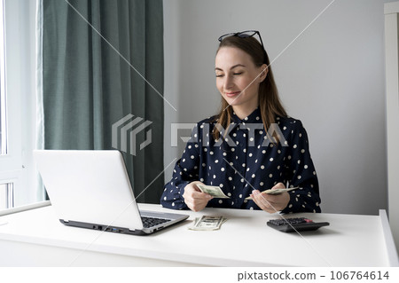 A young woman in a shirt, counting and handing out cash to pay bills at a table, using a laptop 106764614
