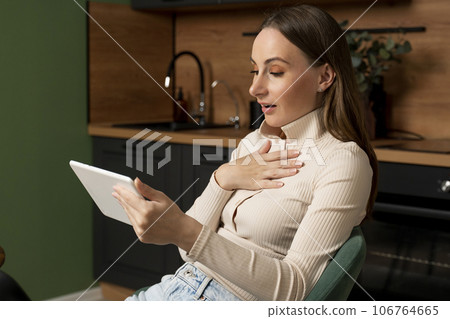 Beautiful brunette woman uses a tablet, enjoys winning an online lottery while sitting in a chair in the kitchen Beautiful brunette woman uses a tablet, enjoys winning an online lottery while sitting in a chair in the kitchen 106764665