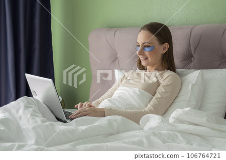 Woman with patches on her face is lying at home on a bed and using a laptop 106764721