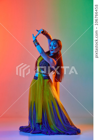 Following asian tradition. Woman in indian dress, makeup and accessories dancing against gradient studio background in neon light Following asian tradition. Woman in indian dress, makeup and accessories dancing against gradient studio background in neon light 106766458