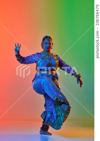 Beautiful indian woman dancing traditional dance in elegant blue dress against gradient studio background in neon light 106766479