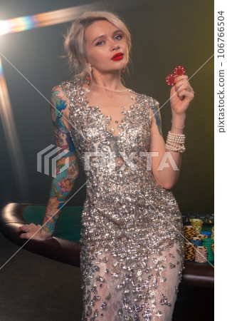 Blonde woman with a beautiful hairstyle and perfect make-up is posing with red gambling chips in her hands. Casino, poker. 106766504