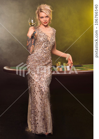 Blonde woman with a beautiful hairstyle and perfect make-up is posing with a glass of champagne in her hand. Casino, poker. Blonde woman with a beautiful hairstyle and perfect make-up is posing with a glass of champagne in her hand. Casino, poker. 106766540