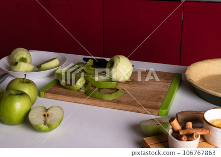 One green pleeling apple on a cutting board and knife on a table with ingredients for apple pie One green pleeling apple on a cutting board and knife on a table with ingredients for apple pie 106767863