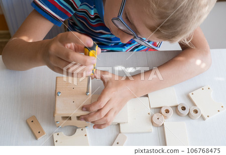 An 8-year-old boy assembles a wooden constructor by himself with a screwdriver. Close-up hands. 106768475