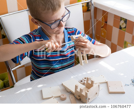 Portrait of a 7-8 year old boy with a screwdriver, carefully assembling a wooden car, sitting at the kitchen table. hands close up. 106768477