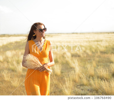 Beauty girl outdoors enjoying nature. A beautiful girl in an orange dress, walks across the field with a bouquet of feather grass, across the steppe, sunlight Beauty girl outdoors enjoying nature. A beautiful girl in an orange dress, walks across the field with a bouquet of feather grass, across the steppe, sunlight 106768490