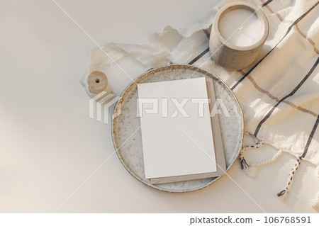 Cozy Christmas or autumn stationery. Closeup of blank greeting card, invitation mockup. Checkered plaid blanket. Candle in concrete cup on white table in sunlight. Flatlay, top view. Copy space. Cozy Christmas or autumn stationery. Closeup of blank greeting card, invitation mockup. Checkered plaid blanket. Candle in concrete cup on white table in sunlight. Flatlay, top view. Copy space. 106768591