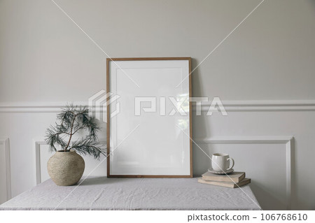 Moody Christmas Scandinavian interior. Minimal winter artistic composition. Blank vertical wooden picture frame mockup, cup of coffee on table. Pine tree branches, old clay vase. White wall background 106768610