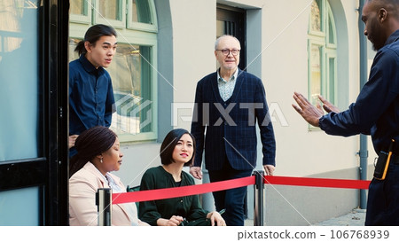 Crowd of people outdoors in line, gathering in the morning and wait to open clothing store door. Anxious customers at entrance on black friday, security guard trying to calm clients. Crowd of people outdoors in line, gathering in the morning and wait to open clothing store door. Anxious customers at entrance on black friday, security guard trying to calm clients. 106768939