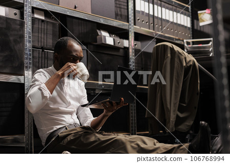Private investigator drinking coffee and using laptop on floor in dark agency office. Police detective analyzing crime case on computer, searching crime case solving solution at night 106768994