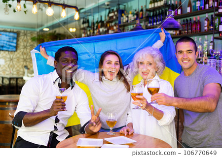 Happy man and woman sport fans holding flag of Ukraine, celebrating and drinking alcoholic drinks in pub, man showing victory sign Happy man and woman sport fans holding flag of Ukraine, celebrating and drinking alcoholic drinks in pub, man showing victory sign 106770649
