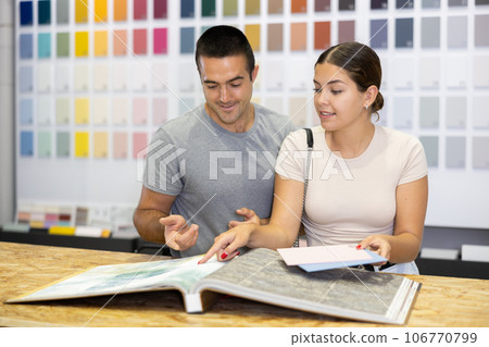 In store, couple buyer look at product catalog and choose color of paint, wall covering in apartment 106770799