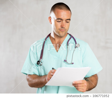 Portrait of male doctor in medical gown with stethoscope making notes in patient medical history Portrait of male doctor in medical gown with stethoscope making notes in patient medical history 106771052