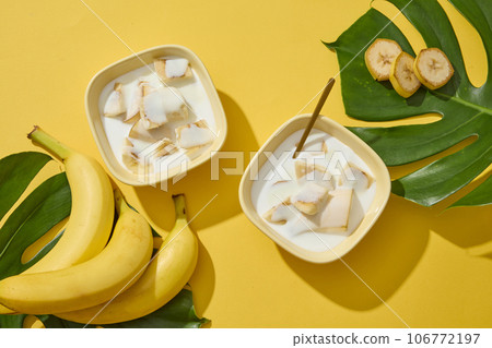 Top view of few bananas and bowls of yogurt and banana slices. Yellow background with tropical leaves. Banana (Musaceae) is a great natural moisturizer Top view of few bananas and bowls of yogurt and banana slices. Yellow background with tropical leaves. Banana (Musaceae) is a great natural moisturizer 106772197