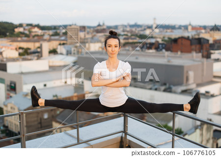 Sportswoman doing side split on roof handrails at sunset Sportswoman doing side split on roof handrails at sunset 106776105
