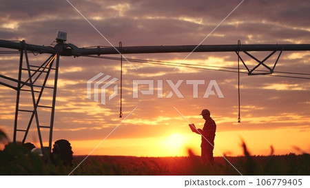 silhouette agriculture. a male farmer works on a laptop in a field with green corn sprouts. corn is watered by irrigation sunlight machine. irrigation agriculture business concept silhouette agriculture. a male farmer works on a laptop in a field with green corn sprouts. corn is watered by irrigation sunlight machine. irrigation agriculture business concept 106779405