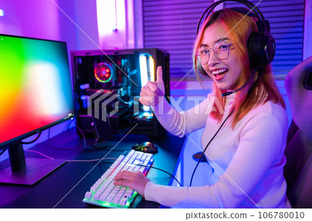 Gamer playing online game wear gaming headphones looking to camera expressing success with game giving thumbs up sign, Smiling woman live stream she play video game at home neon lights living room Gamer playing online game wear gaming headphones looking to camera expressing success with game giving thumbs up sign, Smiling woman live stream she play video game at home neon lights living room 106780010