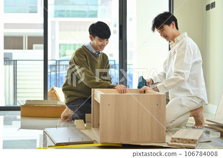 Cheerful young gay male couple using electric drill tool to assembling furniture in their new home Cheerful young gay male couple using electric drill tool to assembling furniture in their new home 106784008
