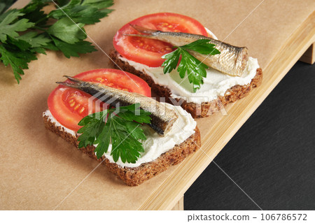 Sandwiches with sprats, tomatoes and parsley on wooden serving tray on black stone background. Appetizer, seafood, front view 106786572