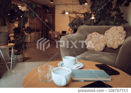 Coffee cups and book on wooden table. Armchair with cushions and table with coffee cups in cafe. Breakfast table in restaurant. Modern urban lifestyle. Morning drinks. Coffee break.  106787553