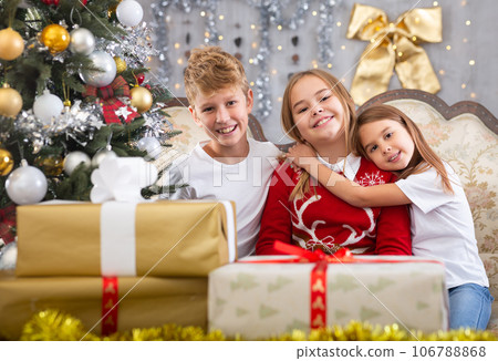 Happy brother with two sisters are sitting on sofa together with gift boxes next to the new year tree Happy brother with two sisters are sitting on sofa together with gift boxes next to the new year tree 106788868
