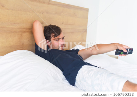 Man watching TV and holding a TV remote on a bed with fan Man watching TV and holding a TV remote on a bed with fan 106790338