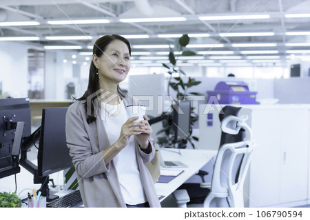 Portrait of a senior business woman sitting on a desk in the office and holding a tumbler Portrait of a senior business woman sitting on a desk in the office and holding a tumbler 106790594