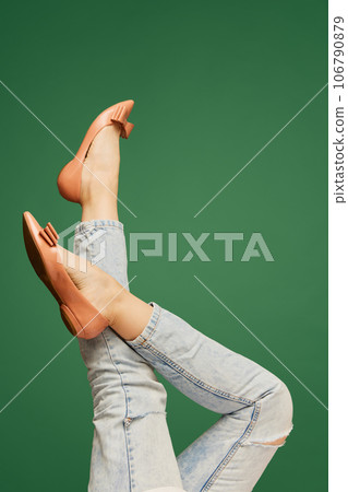 Female slender legs wearing beige flats and casual jeans over green background. Comfort 106790879