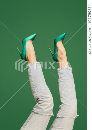 Female slender legs wearing stylish green heeled shoes and casual jeans over green background. Comfort 106790884