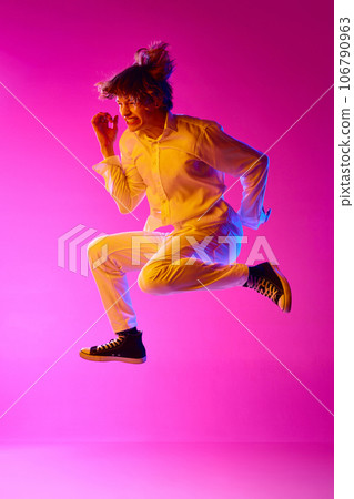 Young guy in casual stylish clothes emotionally jumping against pink studio background in neon lights. Success 106790963