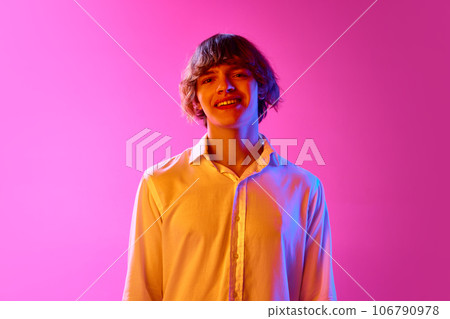Portrait of young guy in white shirt looking at camera and smiling against pink studio background in neon lights 106790978