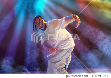 Rap musician. Young artistic man with dreads performing on stage over multicolored neon lights. Concert at night club 106791037