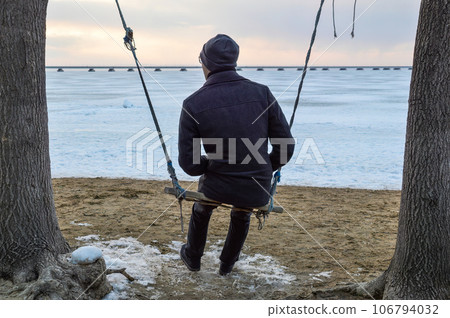 Adult man in a coat swings on a swing and looks thoughtfully at the sea on a cold evening 106794032