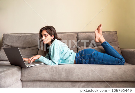 Young woman lying on couch and looking at the laptop at home. Online dating concept. she looking online browsing potential partner profiles. Legs are crossed up under the sofa Young woman lying on couch and looking at the laptop at home. Online dating concept. she looking online browsing potential partner profiles. Legs are crossed up under the sofa 106795449