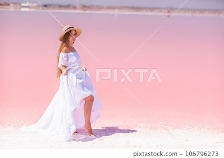 Woman in pink salt lake. She in a white dress and hat enjoys the scenic view of a pink salt lake as she walks along the white, salty shore, creating a lasting memory. 106796273