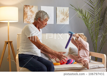 Elderly man, dressed in white t-shirt, sitting on the couch, happily engaging in imaginative play with his two-year-old granddaughter, building colorful pyramid tower together. 106797431