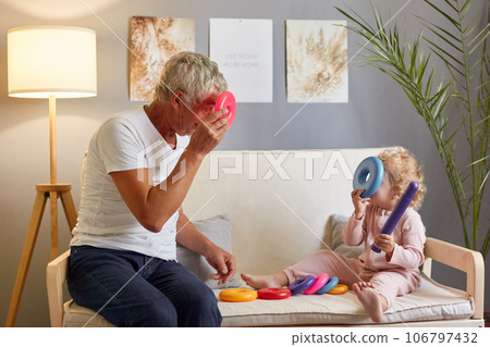 Gray haired mature man wearing white T-shirt sitting on sofa with his baby granddaughter in living room at home connects with his two-year-old granddaughter using colorful pyramid. 106797432