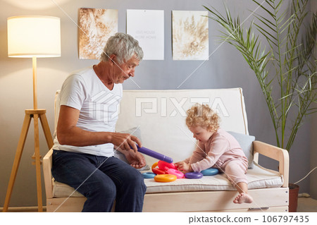 Positive satisfied gray haired senior man wearing white T-shirt sitting on sofa with his baby granddaughter playing with colorful pyramid in living room at home interior. Positive satisfied gray haired senior man wearing white T-shirt sitting on sofa with his baby granddaughter playing with colorful pyramid in living room at home interior. 106797435