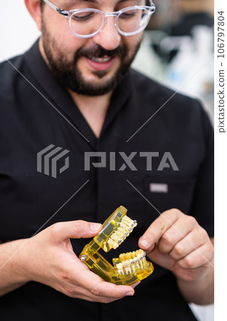 Positive bearded doctor in black uniform explaining dental braces work showing artificial plastic human jaws consulting patient about orthodontic cases in clinic closeup 106797804