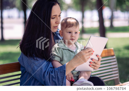 Careful woman in casual outfit showing development cartoons to little son via tablet sitting on bench in urban garden mother using device with baby child in park 106798784