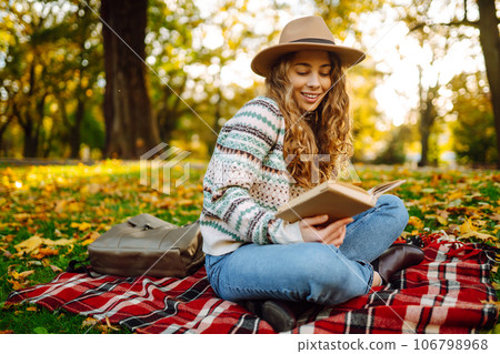 Beautiful curly woman in hat on mat with book in autumn park. Relaxation, solitude with nature. 106798968