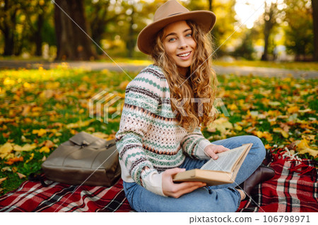 Beautiful curly woman in hat on mat with book in autumn park. Relaxation, solitude with nature. 106798971