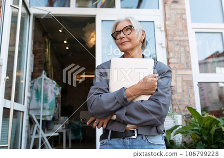 stylish mature woman with gray hair and glasses dressed in a gray jacket holds a laptop for work and stylish mature woman with gray hair and glasses dressed in a gray jacket holds a laptop for work and 106799228