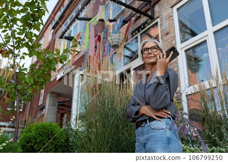 a well-groomed senior woman with gray hair and glasses is dressed in a gray jacket and jeans 106799250