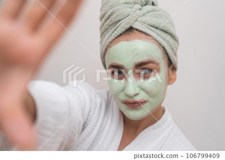 Glowing Self-Portrait: Donning a bathrobe, she grins at the camera, clay mask enhancing her selfie with radiant joy. Glowing Self-Portrait: Donning a bathrobe, she grins at the camera, clay mask enhancing her selfie with radiant joy. 106799409