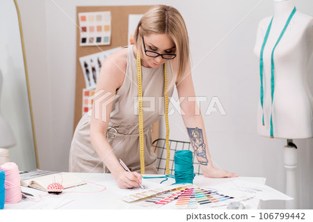Designer with tape measure on neck draws sketch at desk with tailor supplies in sewing atelier dressmaker creates glamorous outfit models working in tailor studio 106799442